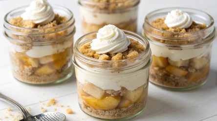 Vier Gläser mit Apple Crumble im Glas´auf hellem Untergrund. Links im Bild liegt ein Löffel.