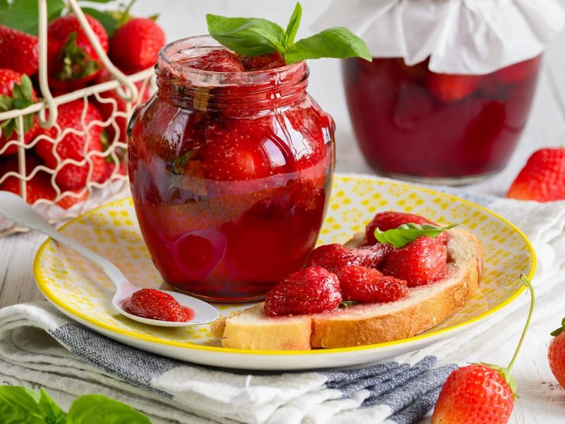 Ein offenes Glas Erdbeer-Basilikum-Marmelade mit Erdbeeren und Basilikumblättern drum herum.