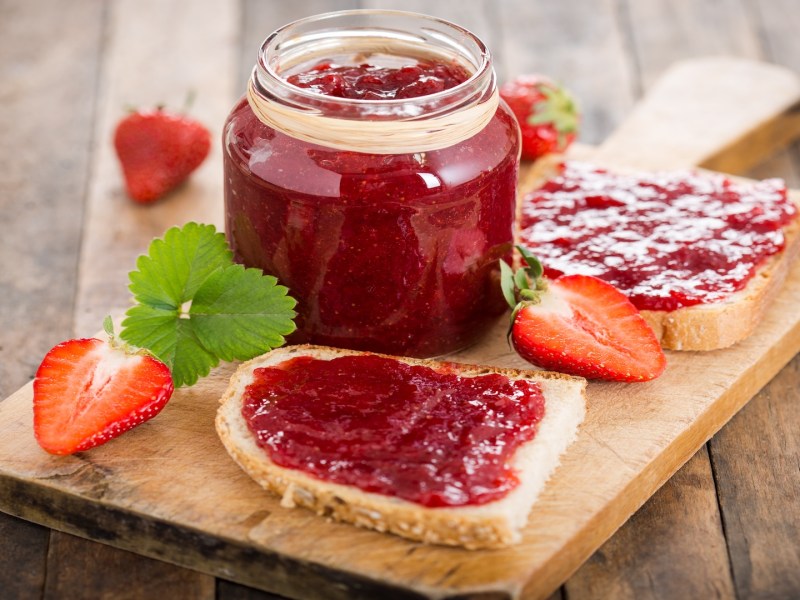 Ein Glas Erdbeer-Vanille-Marmelade auf einem Brett, auf dem auch zwei Brotscheiben mit Marmelade sowie ein paar Erdbeeren liegen.