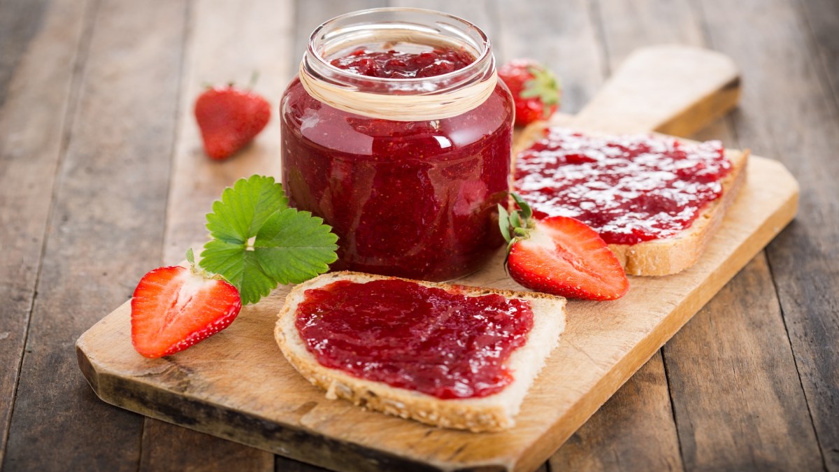 Ein Glas Erdbeer-Vanille-Marmelade auf einem Brett, auf dem auch zwei Brotscheiben mit Marmelade sowie ein paar Erdbeeren liegen.
