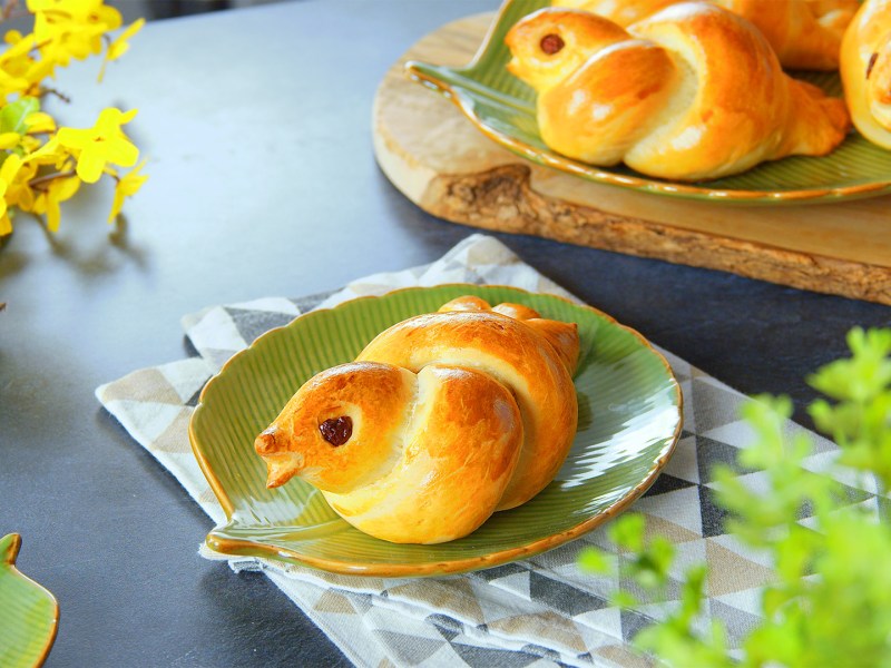 Eine Hefezopftaube liegt auf einem grünen, blattförmigen Teller. Im Hintergrund liegen weitere Hefezopftauben. Der Teller steht auf einem Küchentuch. Im Vordergrund sowie im Hintergrund ist etwas Pflanzendeko.