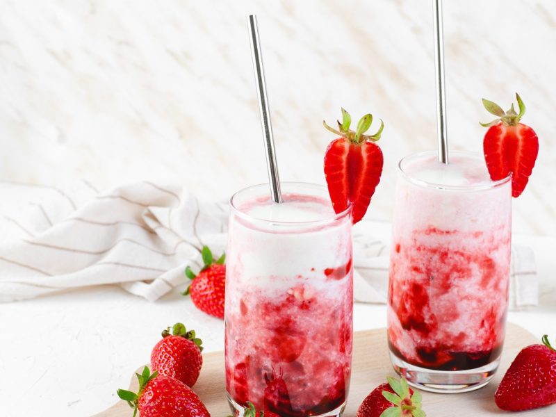 Zwei Gläser mit koreanischer Erdbeermilch, die jeweils mit einer Erdbeere am Glasrand garniert und mit einem Strohhalm serviert werden. Ringsum liegen frische Erdbeeren und man kann eine weiße Schale mit Erdbeeren erkennen im Hintergrund.