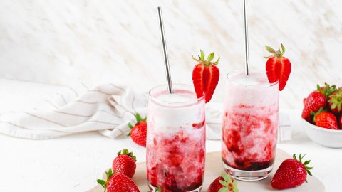 Zwei Gläser mit koreanischer Erdbeermilch, die jeweils mit einer Erdbeere am Glasrand garniert und mit einem Strohhalm serviert werden. Ringsum liegen frische Erdbeeren und man kann eine weiße Schale mit Erdbeeren erkennen im Hintergrund.