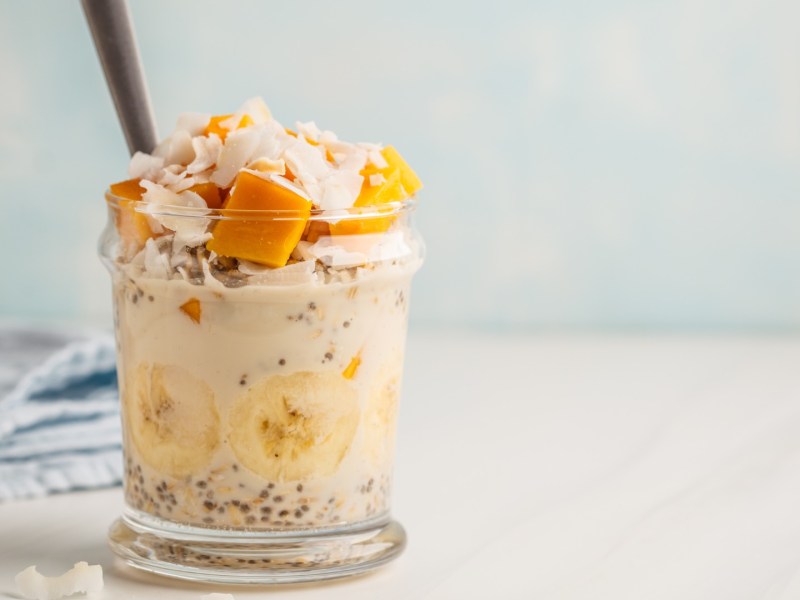 Mango-Kokos-Overnight-Oats im Glas geschichtet und serviert mit einem Strohhalm.