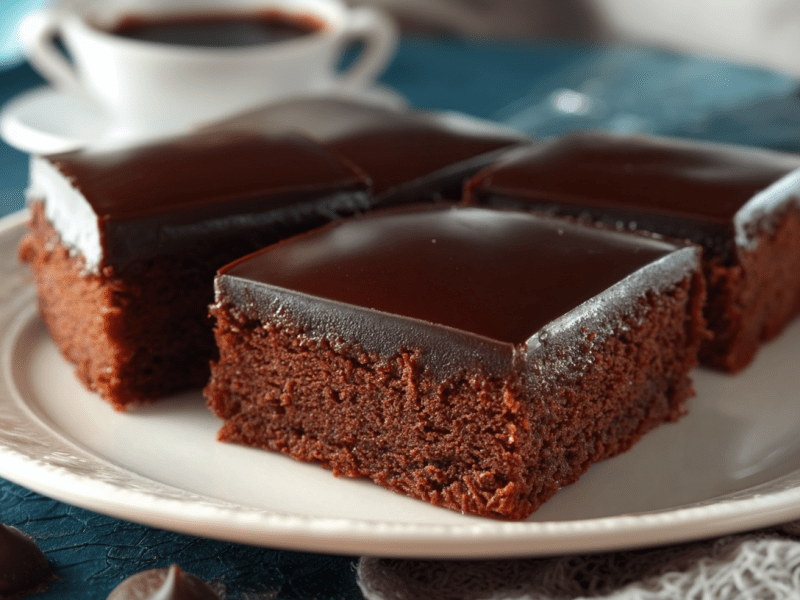Vier Stücke Sachertorte vom Blech liegen auf einem weißen Teller. Im Hintergrund steht eine Kaffeetasse. Um den Teller verteilt liegen Schokodrops.