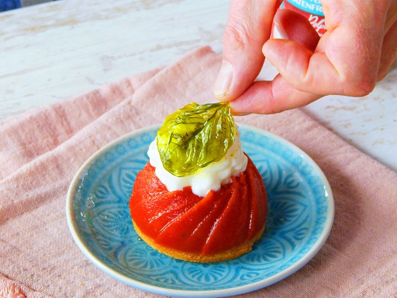 Ein Teller mit Tomaten-Panna-cotta, die mit einem Mozzarella-Schaum und einem frittierten Basilikumblatt garniert ist. Man sieht, wie eine Hand, das Basilikumblatt auf den Mozzarella-Schaum setzt. Der Teller steht auf einem rosafarbenen Tuch.