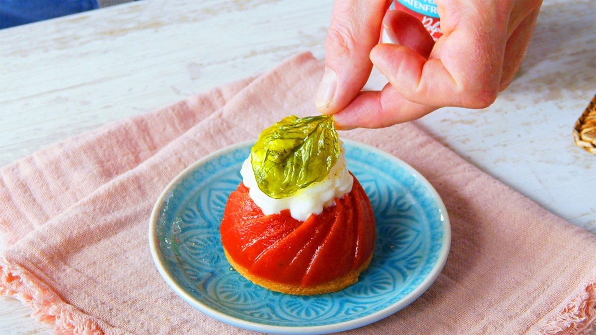 Ein Teller mit Tomaten-Panna-cotta, die mit einem Mozzarella-Schaum und einem frittierten Basilikumblatt garniert ist. Man sieht, wie eine Hand, das Basilikumblatt auf den Mozzarella-Schaum setzt. Der Teller steht auf einem rosafarbenen Tuch.