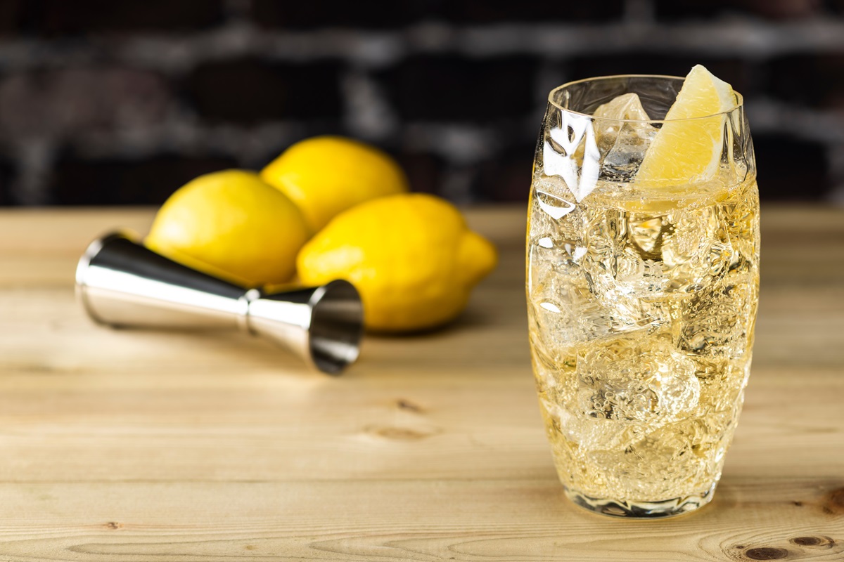 Ein Glas mit Whisky Soda steht auf einem Holztisch. Im Hintergrund liegen drei Zitronen sowie ein Cocktail-Messbecher.