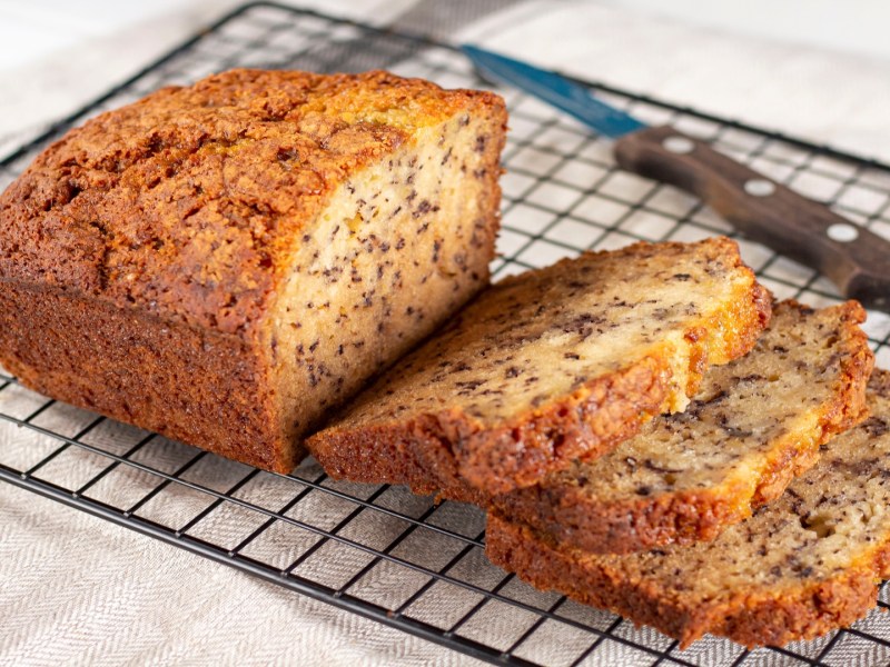 Ein angeschnittenes Zitrone-Bananenbrot mit Mohn auf einem Kuchengitter, daneben ein Messer.