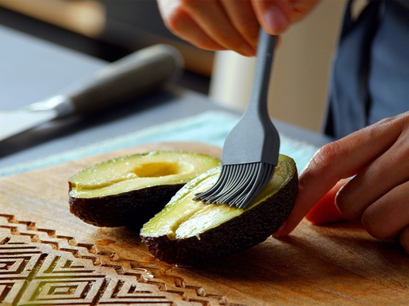 Zwei Acocadohälften auf einem Holzbrett. Sie werden von einer Köchin eingepinselt.