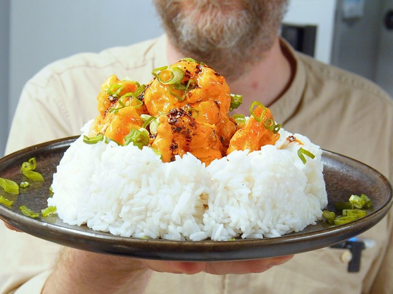 Ein Teller mit einer großen Portion Reis, auf der die Bang Bang Garnelen verteilt sind. Koch Oli hält den Teller in die Kamera.