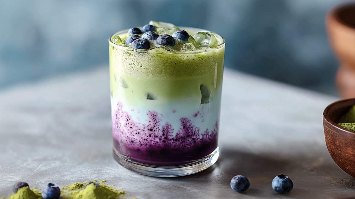 Ein Glas mit Blaubeer-Matcha-Latte. Auf der Arbeitsfläche kann man noch ein Häufchen Matcha-Pulver sowie eine Schale mit Matcha-Pulver erkennen. Außerdem liegen ein paar einzelne Heidelbeeren herum.