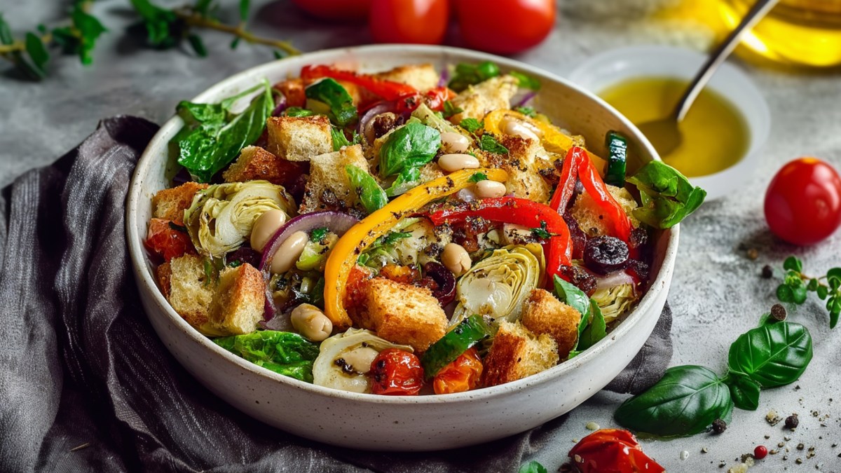 Ein großer Teller Brotsalat mit gerösteter Paprika. Ringsum sind frische Zutaten wie Basilikum und Tomaten zu sehen. Außerdem steht ein Schälchen mit Öl und einem Löffel drin auf der Arbeitsfläche.