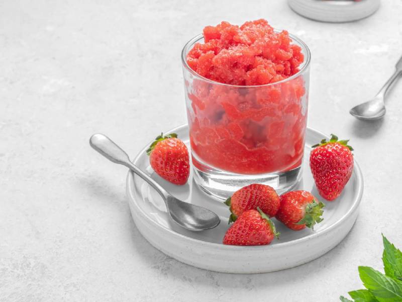 Ein Glas Erdbeer-Granita. Drum herum liegen Erdbeeren und Basilikumblätter.