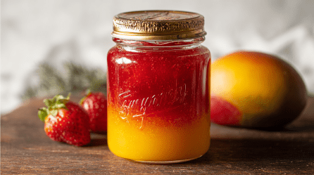 Ein Glas Erdbeer-Mango-Marmelade. Im Hintergrund zwei Erdbeeren und eine Mango.