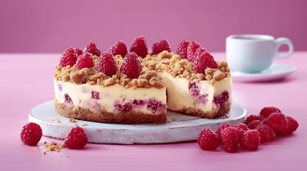 Ein angeschnittener Himbeer-Quark-Kuchen steht auf einer Platte. Davor liegen ein paar Himbeeren. Im Hintergrund eine Kaffeetasse.