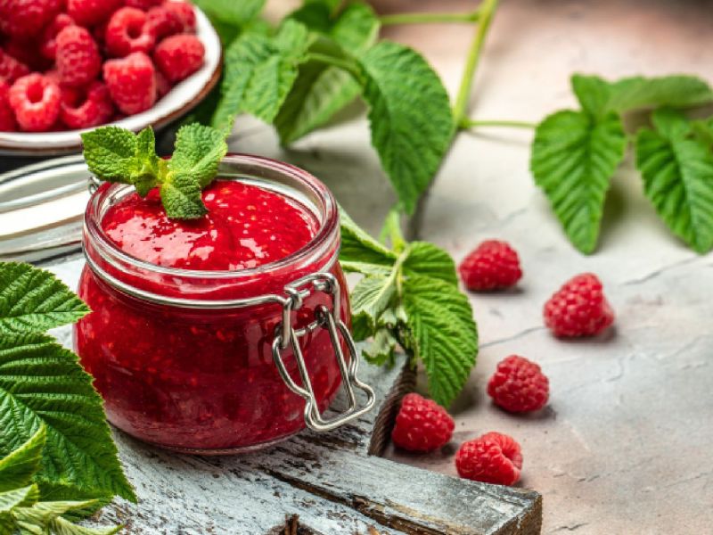 Ein Glas Himbeermarmelade, dahinter eine Schale mit frischen Himbeeren. Drum herum liegen einige Himbeerblätter.