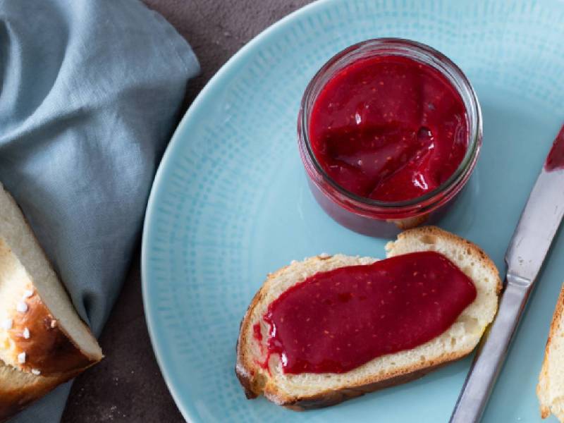 Teller mit zwei Scheiben Brot, bestrichen mit Himbeermarmelade ohne Kerne, Messer und ein Glas Marmelade daneben
