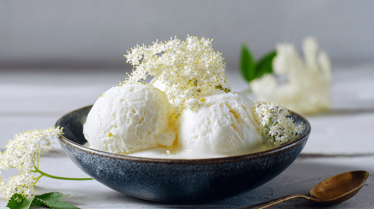 Eine Schale mit drei Kugeln Holunderblüten-Eis, dekoriert mit einigen Holunderblüten.