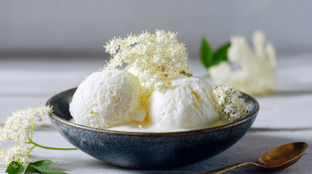 Eine Schale mit drei Kugeln Holunderblüten-Eis, dekoriert mit einigen Holunderblüten.
