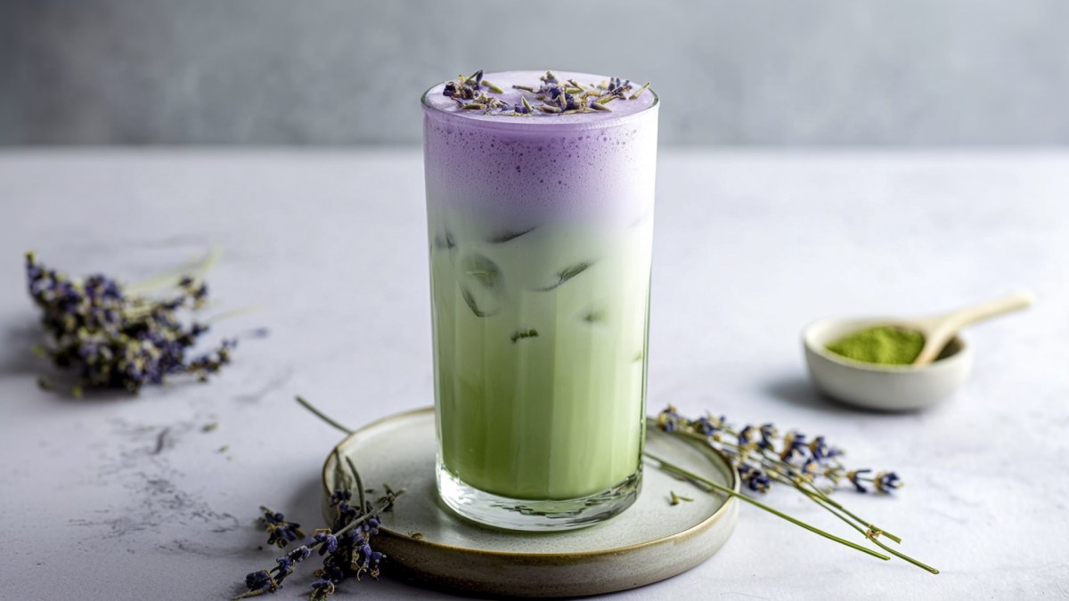 Ein Glas mit Iced-Lavendel-Matcha-Latte, das auf einem Keramikuntersetzer steht. Ringsum liegen einige Zweige Lavendel herum. Außerdem steht eine kleine Schale mit Matcha-Pulver im Hintergrund.