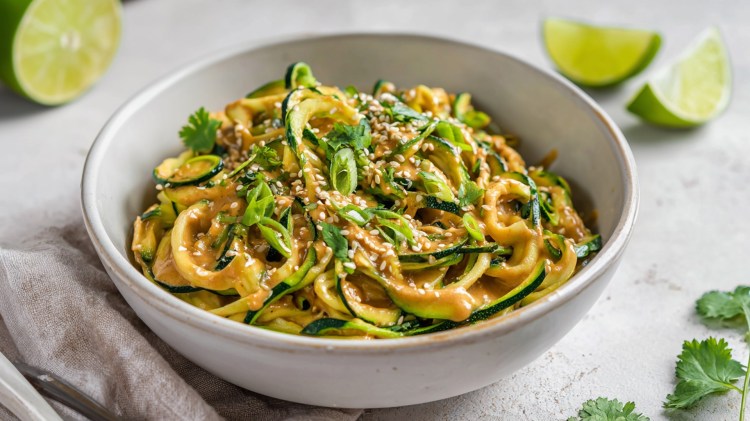 Eine große Schüssel voll kalter Zucchini-Nudeln mit Erdnuss-Sesam-Soße, die mit Sesam sowie Frühlingszwiebeln garniert sind. Um die Schüssel herum liegen Limettenschnitze sowie etwas frischer Koriander. Auch ein Küchenhandtuch ist zu sehen.