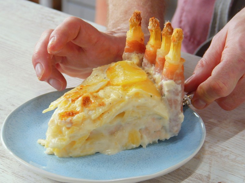 Ein Stück Kartoffel-Spargelauflauf auf einem Teller. Es sind die Hände des Kochs zu sehen.