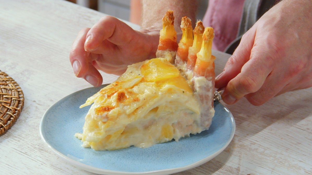 Ein Stück Kartoffel-Spargelauflauf auf einem Teller. Es sind die Hände des Kochs zu sehen.