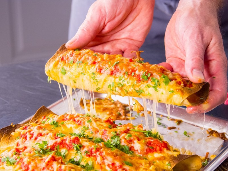 Knusprige Lasagnerollen auf einem Backblech. Eine wird hochgehoben und zieht käsige Fäden.