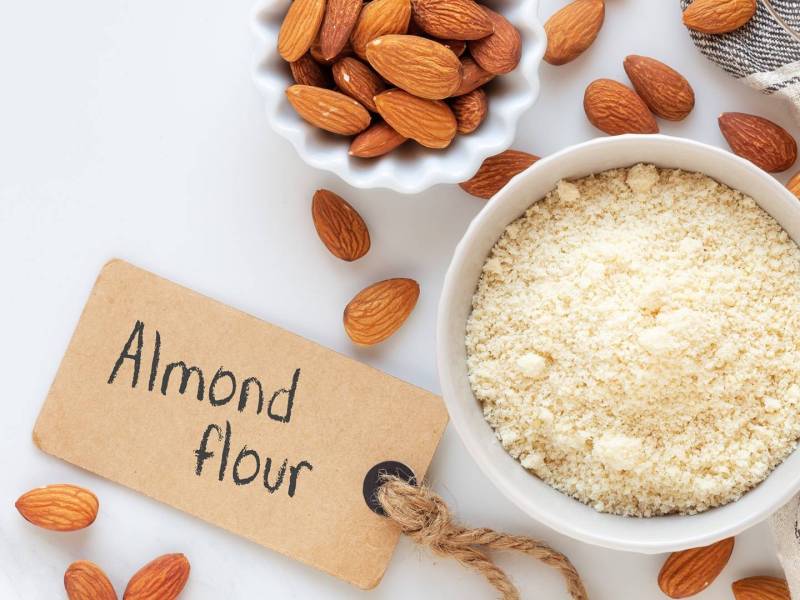 Draufsicht auf eine Schale mit Mandelmehl und eine kleinere Schale mit Mandeln. Drum herum liegen braune Mandeln verteilt. Außerdem ist ein Schild zu sehen, auf dem 