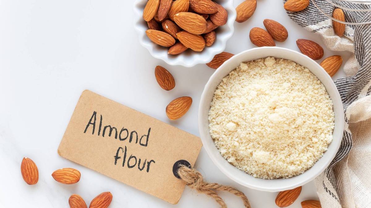 Draufsicht auf eine Schale mit Mandelmehl und eine kleinere Schale mit Mandeln. Drum herum liegen braune Mandeln verteilt. Außerdem ist ein Schild zu sehen, auf dem 