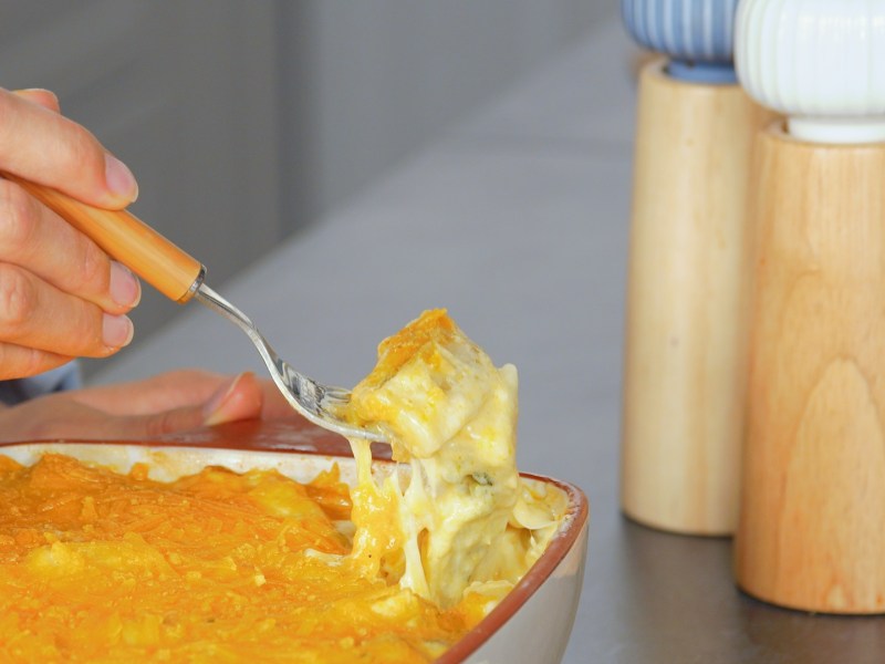 Eine Auflaufform mit Maultaschen-Auflauf mit Spargel, die auf einer Arbeitsfläche steht. Man kann erkennen, wie eine Hand mit einer Gabel etwas aus dem Auslauf herausgabelt. Neben der Form kann man einen Salz- und Pfefferstreuer erkennen.
