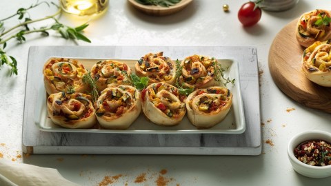 Eine Servierplatte mit einigen mediterranen Gemüseschnecken. Die Platte steht auf einem hellen Brett. Rechts daneben auf einem Holzbrett liegen weitere Gemüseschnecken. Ringsum sind frische Kräuter und Gewürze zu sehen.