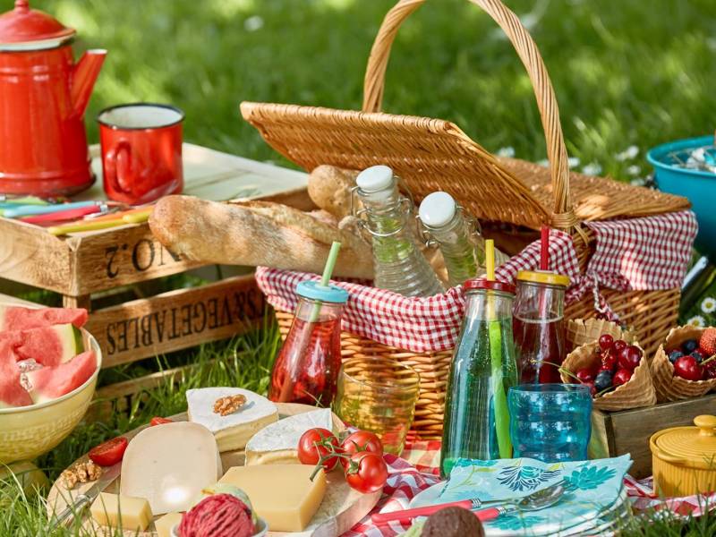 Ein Picknick-Arrangement, dass aus einem Picknickkorb auf einer Wiese besteht. Man kann Glasgefäße mit Getränken, eine Käseplatte, Schüsseln und Waffeln mit Früchten, ein kleinen Grill mit Maiskolben sowie Baguette, das im Picknickkorb ist, erkennen. Außerdem steht eine kleine Weinkiste als Tisch daneben, auf der eine rote Kaffeekanne und ein roter Kaffeebecher steht.