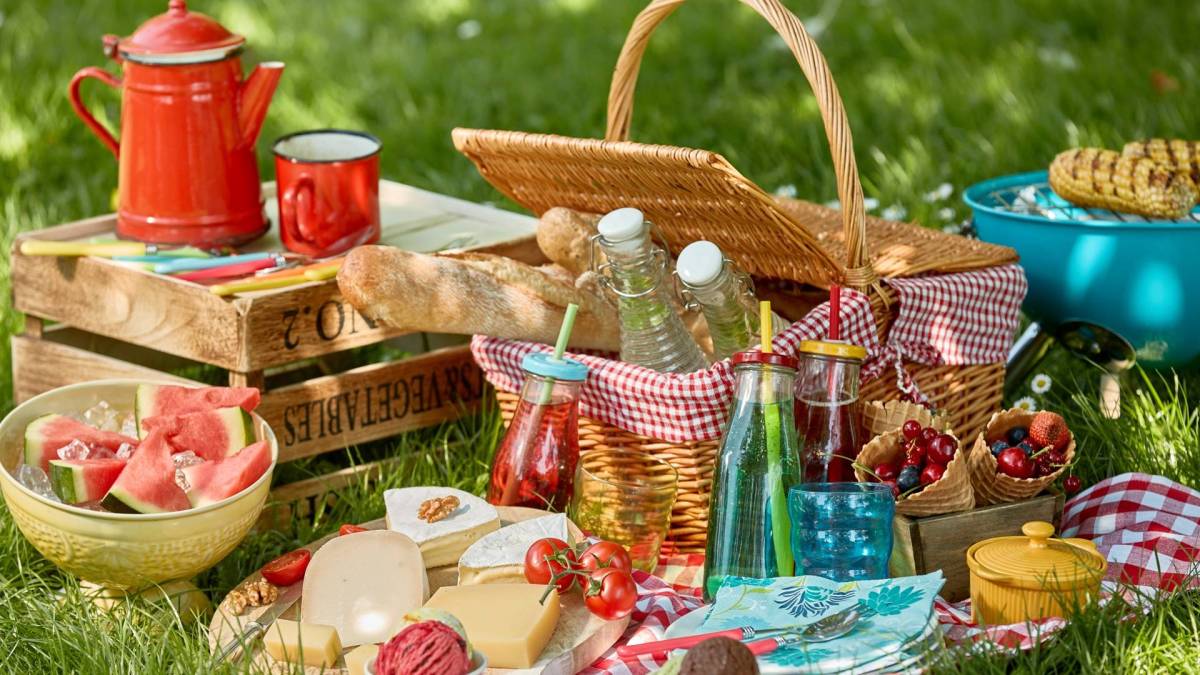 Ein Picknick-Arrangement, dass aus einem Picknickkorb auf einer Wiese besteht. Man kann Glasgefäße mit Getränken, eine Käseplatte, Schüsseln und Waffeln mit Früchten, ein kleinen Grill mit Maiskolben sowie Baguette, das im Picknickkorb ist, erkennen. Außerdem steht eine kleine Weinkiste als Tisch daneben, auf der eine rote Kaffeekanne und ein roter Kaffeebecher steht.