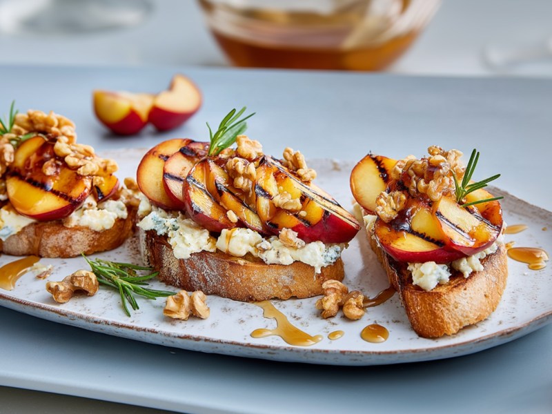 Ein Servierteller mit einigen Scheiben Nektarinen-Bruschetta. Außerdem liegen einzelne Nektarinenstücke und ein frischer Zweig Rosmarin herum.