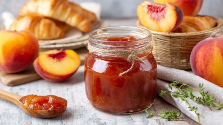 Ein Glas mit selbst gemachter Nektarinen-Thymian-Marmelade. Daneben liegt ein Holzlöffel mit Marmelade drauf. Im Hintergrund kann man einen Teller mit Croissants erkennen. Außerdem liegen Ganze Nektarinen und Nektarinenschnitze herum genauso wie etwas frischer Thymian.