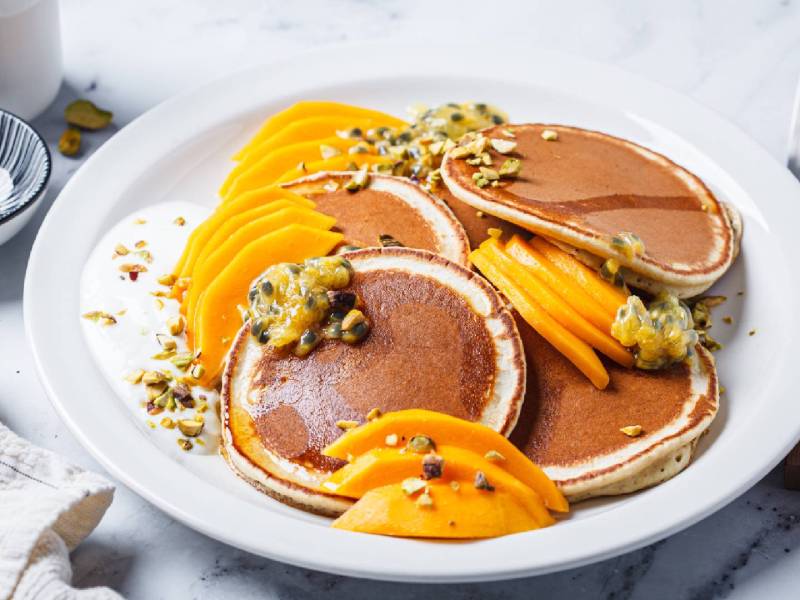 Pancakes mit Mango und Passionsfrucht auf einem weißen Teller, garniert mit gehackten Pistazien.