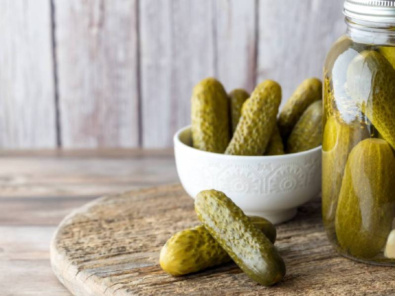 Ein Glas eingelegte Salz-Dill-Gurken neben einer Schüssel mit ebendiesen Gurken.