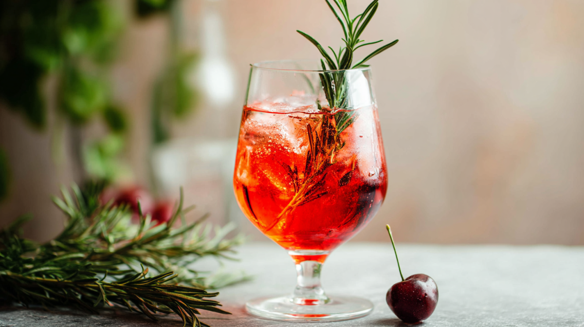 Ein Glas Savoia Spritz, garniert mit Rosmarin und einer Kirsche.