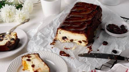 Ein weißes Backpapier auf dem ein angeschnittener ukrainischer Käsekuchen liegt. Auf Tellern, die daneben stehen, liegen Stücke des Kuchens. Außerdem kann man ein paar weiße Blumen erkennen und es steht eine Schale mit Rosinen da. Ein Messer liegt beim Kuchen.