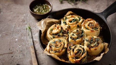 Eine tiefe Gusseisen-Pfanne mit veganen Spinat-Schnecken. Daneben liegt ein dünnes Messer. Außerdem steht eine kleine Schale mit flüssiger Knoblauchbutter daneben.