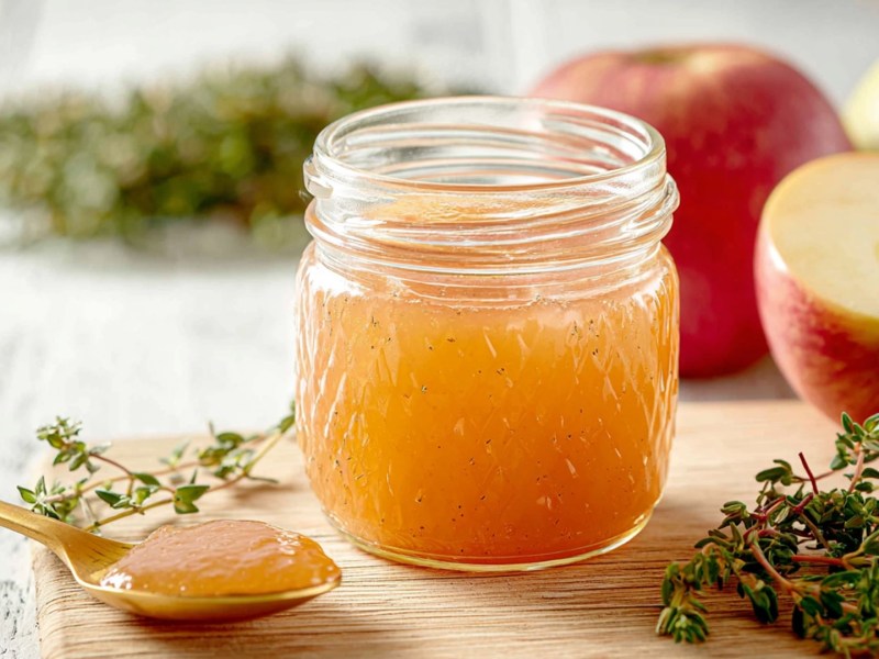Ein Glas Apfel-Thymian-Marmelade, das auf einem Holzbrett steht. Daneben liegt eine Apfelhälfte sowie ganze Äpfel. Auch frischer Thymian sowie ein Löffel mit Marmelade sind zu sehen.