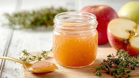 Ein Glas Apfel-Thymian-Marmelade, das auf einem Holzbrett steht. Daneben liegt eine Apfelhälfte sowie ganze Äpfel. Auch frischer Thymian sowie ein Löffel mit Marmelade sind zu sehen.