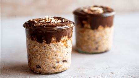 Zwei Gläser Bananenbrot Overnight Oats stehen auf neutralem Untergrund. Sie sind mit einer Schicht Schokolade bedeckt.