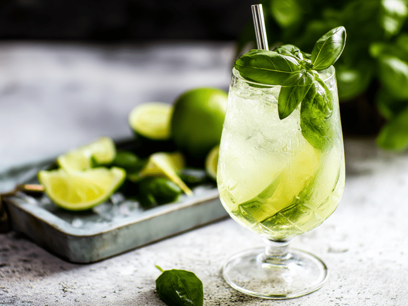 Ein Glas Basilikum Spritz vor einem Tablett mit Limetten und etwas Basilikum.