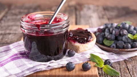 Ein offenes Glas Blaubeer-Limetten-Konfitüre, in dem ein Löffel steckt. Daneben frische Blaubeeren und ein Brot mit der Konfitüre bestrichen.