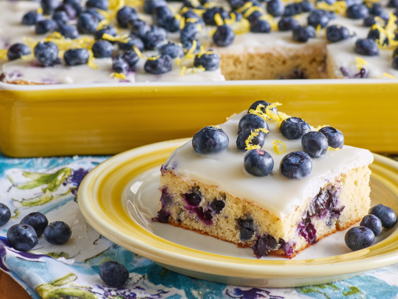 Ein Stück Blaubeer-Zitronen-Kuchen vom Blech. Im Hintergrund ist der restliche Kuchen.
