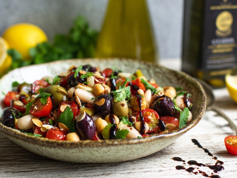 Ein Teller mit Bohnen-Oliven-Salat. Im Hintergrund liegen Zitronen, frische Kräuter und es stehen Flaschen mit Olivenöl.