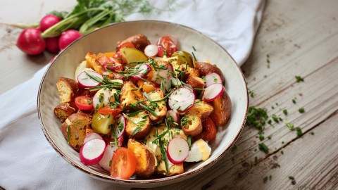 Ein Teller mit Brezelsalat, der auf einem Tisch steht. Im Hintergrund kann man einen kleinen Bund Radieschen erkennen. Rechts neben dem Teller liegen frisch gehackte Kräuter verstreut.